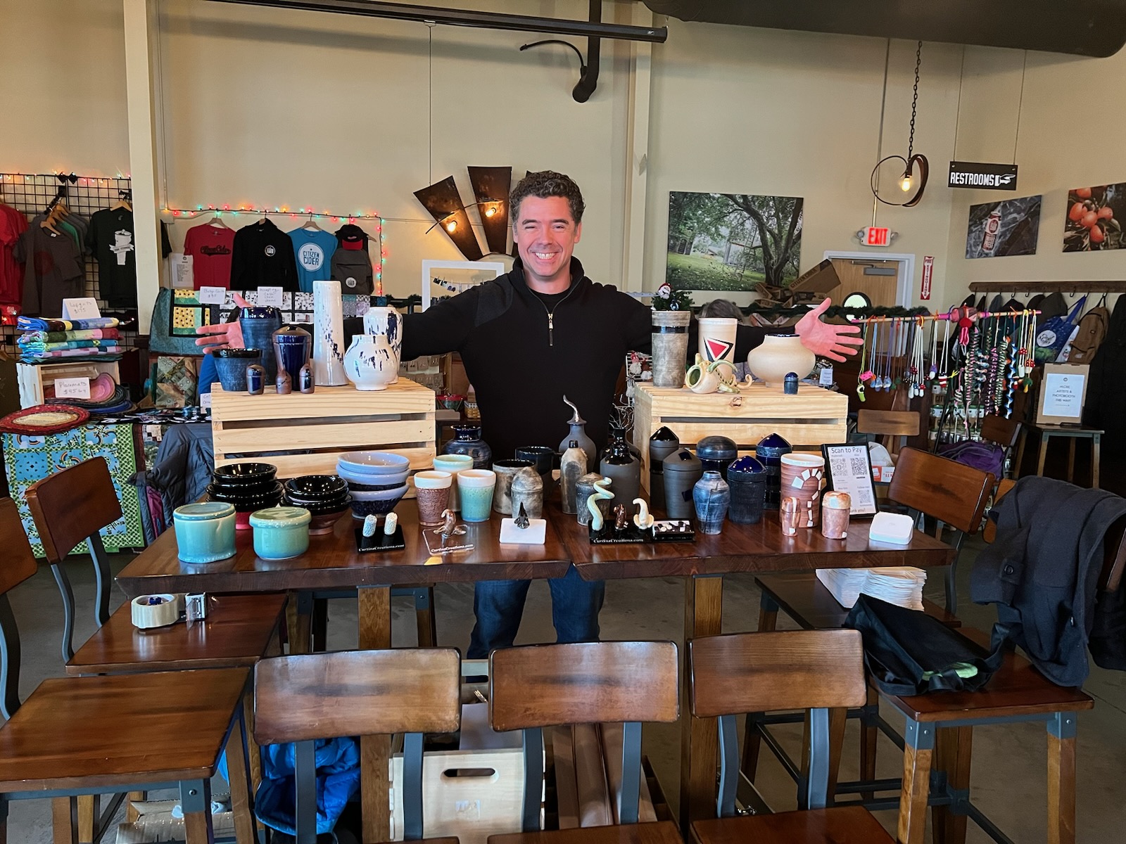 Tim behind a market table of his pottery, arms spread wide, with mugs, bowls, vases and lidded jars on wooden crates